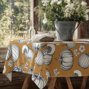 Op de afbeelding: Een mosterdgeel tafelkleed met een herhalend zwart-wit lijnpatroon van pompoenen, pompoenplakken en kleine bloemen. De stof is gedrapeerd over een houten tafel, met een gegalvaniseerde metalen emmer met witte bloemen erop.