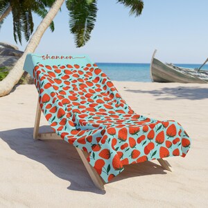 May include: A light blue beach towel with a red strawberry pattern and the name "shannon" printed on it. The towel is draped over a wooden beach chair.