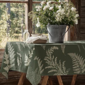Puede incluir: Un mantel verde con un diseño de helechos cubre una mesa de madera. Un cubo de metal galvanizado lleno de flores blancas está sobre la mesa, junto con una pila de platos, una botella de vidrio y un paño blanco. La escena está cerca de una ventana.