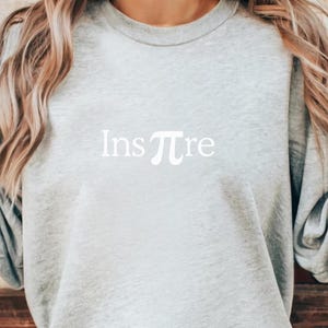 以下が含まれることがあります： 白で「Inspπre」とプリントされたライトグレーのスウェットシャツ。デザインは「Ins」の後に数学記号のπ、そして「re」が続きます。スウェットシャツはカジュアルなスタイルです。