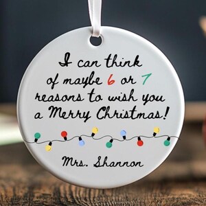 May include: A white ceramic Christmas ornament with the text "I can think of maybe 6 or 7 reasons to wish you a Merry Christmas!" and "Mrs. Shannon". The ornament has a string of colorful lights and a white ribbon for hanging.