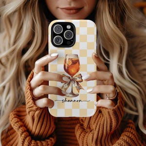 May include: A phone case with a yellow and white checkered pattern. The case features a graphic of a cocktail in a glass with a bow. The name "shannon" is written in cursive below the graphic.