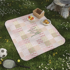 May include: A pink and yellow patchwork picnic blanket with a floral pattern and the letter "M" embroidered in the center.