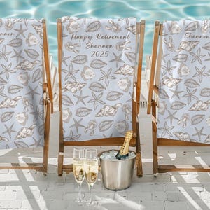 May include: Two blue and white beach chairs with a seashell and starfish pattern. The chair in the middle has the text "Happy Retirement Shannon 2025" printed on it. A bottle of champagne and two champagne flutes are in front of the chairs.