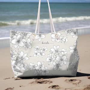May include: A large tote bag with a floral design and the word "bride" in script. The bag features a light beige background with black and white flower illustrations. The bag has white rope handles and is sitting on a sandy beach.