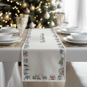 Peut inclure: Chemin de table blanc avec une bordure de maisons et d'arbres d'hiver illustrés. Le texte "The Smith Family" est imprimé en noir. Le chemin de table est posé sur une table avec de la vaisselle blanche et des verres dorés.