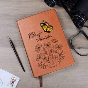 Puede incluir: Un diario de cuero marr&oacute;n con las palabras "Change is Beautiful" y un gr&aacute;fico de mariposa. El diario tambi&eacute;n presenta un dise&ntilde;o floral. Tambi&eacute;n se ven un bol&iacute;grafo, unos prism&aacute;ticos y un sobre.