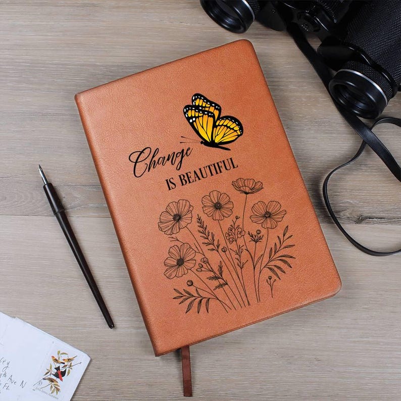 Puede incluir: Un diario de cuero marr&oacute;n con las palabras "Change is Beautiful" y un gr&aacute;fico de mariposa. El diario tambi&eacute;n presenta un dise&ntilde;o floral. Tambi&eacute;n se ven un bol&iacute;grafo, unos prism&aacute;ticos y un sobre.