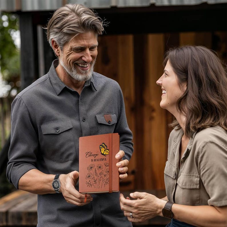 Puede incluir: Un diario marr&oacute;n con la frase "Change is Beautiful" y un dise&ntilde;o de mariposa. El diario tiene una correa de cuero e ilustraciones florales. Lo sostiene una persona con una camisa gris.