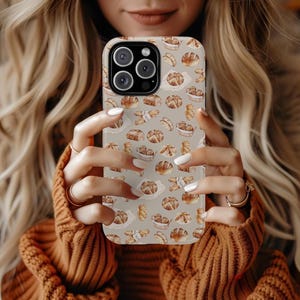 May include: A phone case featuring a repeating pattern of various baked goods, including bread loaves and rolls, on a light beige background. The case has a glossy finish and a camera cutout.