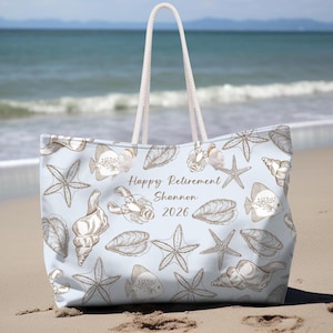May include: Light blue beach tote bag with a repeating pattern of seashells, starfish, and fish. The bag has white rope handles and the text "Happy Retirement Shannon 2026" printed on the front.