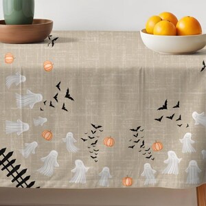 K&ouml;nnte beinhalten: Eine beige Tischdecke mit einem wiederkehrenden Muster aus wei&szlig;en Geistern, schwarzen Flederm&auml;usen und orangefarbenen K&uuml;rbissen. Ein schwarzes Lattenzaun-Design ist in der unteren linken Ecke aufgedruckt. Der Stoff hat eine feine Gitterstruktur.