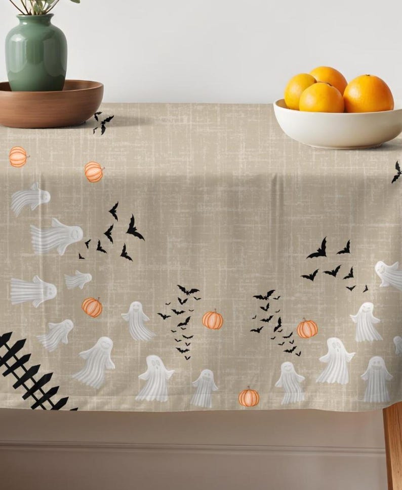 K&ouml;nnte beinhalten: Eine beige Tischdecke mit einem wiederkehrenden Muster aus wei&szlig;en Geistern, schwarzen Flederm&auml;usen und orangefarbenen K&uuml;rbissen. Ein schwarzes Lattenzaun-Design ist in der unteren linken Ecke aufgedruckt. Der Stoff hat eine feine Gitterstruktur.