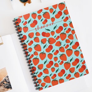 May include: A blue spiral notebook with a red and white strawberry and cherry pattern. The name "shannon" is written in red cursive on the front cover.