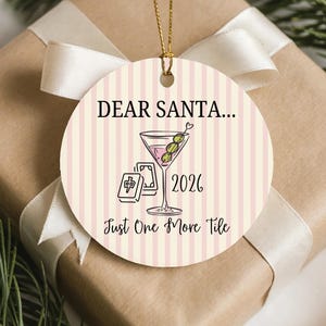 May include: A round ceramic ornament with a light pink and white vertical striped background. It features a martini glass with olives, mahjong tiles, the text DEAR SANTA... 2026, and Just One More Tile. A gold string is attached to the top.