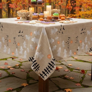 K&ouml;nnte beinhalten: Eine quadratische, beige Tischdecke mit einem wiederkehrenden Halloween-Muster aus wei&szlig;en Geistern, orangefarbenen K&uuml;rbissen, schwarzen Flederm&auml;usen und einer schwarzen Zaunbord&uuml;re am unteren Rand. Der Stoff wirkt strukturiert und bedeckt einen Holztisch.