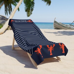 May include: A blue and white striped beach towel with the word "MAINE" and red lobster illustrations. The towel is draped over a wooden beach chair on a sandy beach.