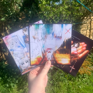 May include: Three square art prints held in a hand, featuring anime-style illustrations and text. One print has the text "If you mean the creator of the world." Another reads "It's not me." The prints have a watercolor aesthetic.