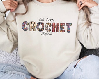May include: A cream-colored sweatshirt with the text "Eat, Sleep, Crochet, Repeat" in black. The word "Crochet" is made up of colorful crocheted squares.