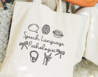 May include: Cream-colored tote bag with black line art illustrations and text. The design includes a speech bubble, brain, lips, bow, ear, hand gesture, and the words "Speech Language Pathologist."