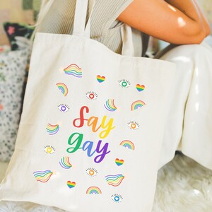 May include: A white canvas tote bag with the phrase "Say Gay" in rainbow letters and rainbow-colored hearts, eyes, and rainbows surrounding the text.