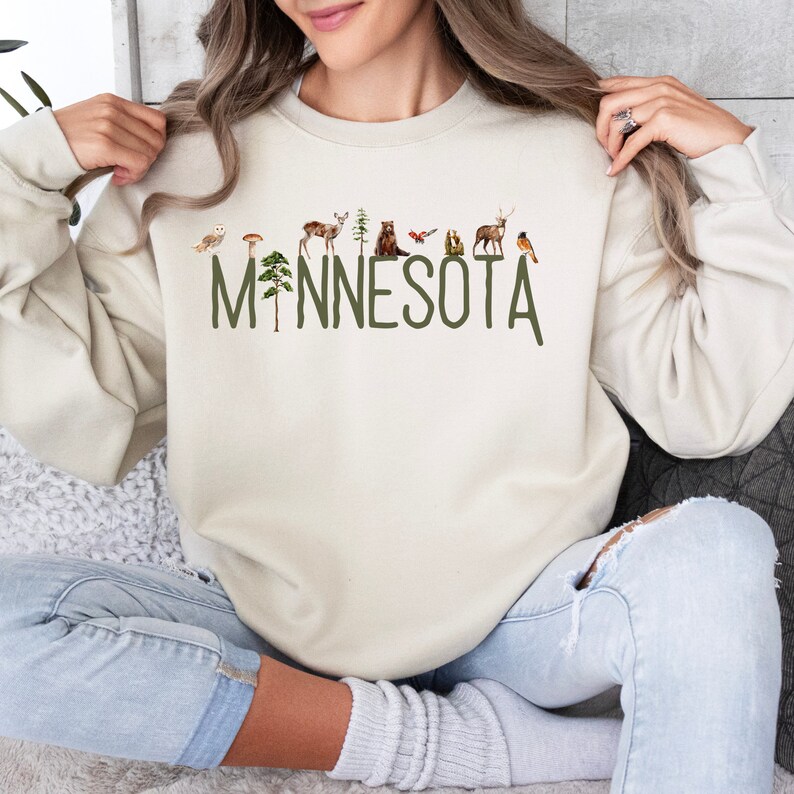 K&ouml;nnte beinhalten: Ein cremefarbener Pullover mit einer gr&uuml;nen Grafik des Wortes "MINNESOTA" mit verschiedenen Waldtieren und Pflanzen.