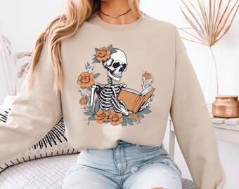 Pode incluir: Camisola bege com um esqueleto lendo um livro, rodeado por flores laranja e folhas verdes. O esqueleto tem uma flor no cabelo. O desenho é impresso na frente da camisola.