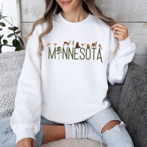 K&ouml;nnte beinhalten: Wei&szlig;er Pullover mit einer gr&uuml;nen Grafik des Wortes "MINNESOTA" mit verschiedenen Waldtieren und Pflanzen.