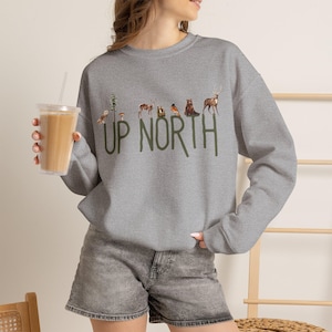 May include: A heather gray crewneck sweatshirt featuring the green text UP NORTH with small watercolor-style illustrations of an owl, a pine tree, a mushroom, a deer, a bear, and a stag positioned above the letters on the front of the garment.