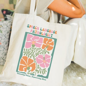 May include: A white canvas tote bag with the text "Speech Language Pathologist" and a floral design. The text "Unlocking Voices, Empowering Lives" is printed below the floral design.