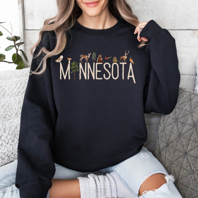 K&ouml;nnte beinhalten: Schwarzer Pullover mit einem wei&szlig;en Aufdruck des Wortes "MINNESOTA" mit einer Reihe kleiner Waldtiere und Pflanzen darunter.