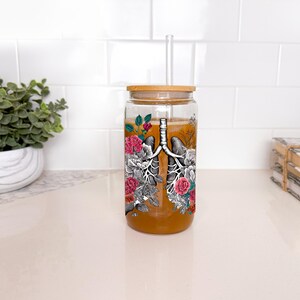 May include: A clear glass reusable water bottle with a bamboo lid and a straw. The bottle has a graphic of a pair of lungs with flowers surrounding them.