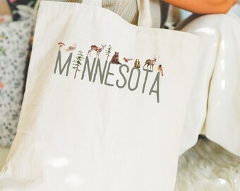 May include: Cream-colored tote bag with the word "MINNESOTA" in gray letters. The word is decorated with illustrations of woodland creatures, including an owl, deer, and bear. The bag has two handles.