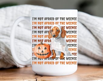 May include: White ceramic mug featuring a dachshund dressed as a ghost next to an orange jack-o'-lantern. The mug has the text "I'M NOT AFRAID OF THE WEENIE" in orange and black. A fun Halloween-themed gift.