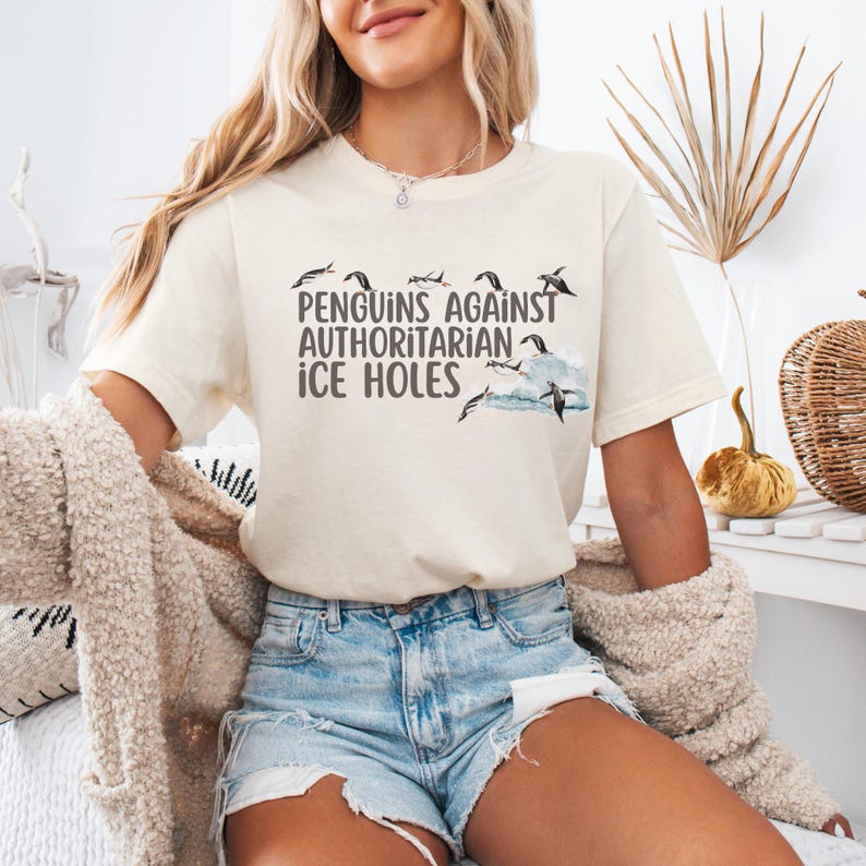 May include: A white t-shirt with a graphic of penguins jumping over waves and the text "Penguins Against Authoritarian Ice Holes".