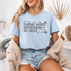 May include: A light blue t-shirt with a graphic of penguins jumping over waves. The text "Penguins Against Authoritarian Ice Holes" is printed on the shirt.