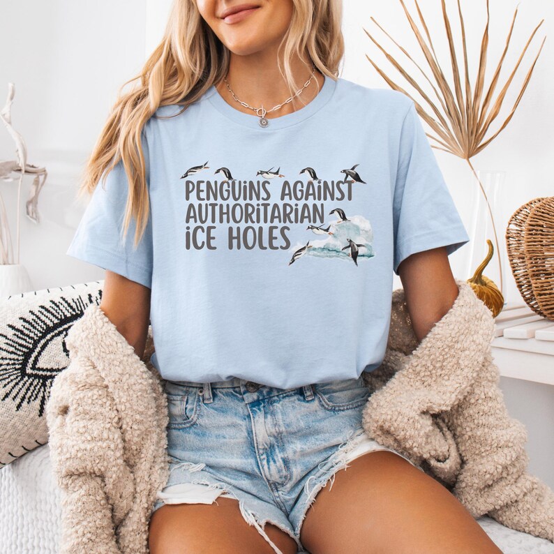 May include: A light blue t-shirt with a graphic of penguins jumping over waves. The text "Penguins Against Authoritarian Ice Holes" is printed on the shirt.
