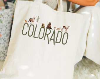 May include: Off-white canvas tote bag featuring the word "COLORADO" in green, with watercolor illustrations of wildlife above the letters. The illustrations include a bird, deer, tree, bear, owl, and deer.