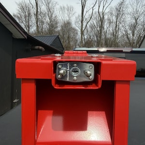 May include: A bright red metal storage box with a rectangular shape. The box has a hinged lid and a silver Master Lock on the front. The background includes bare trees and a cloudy sky.