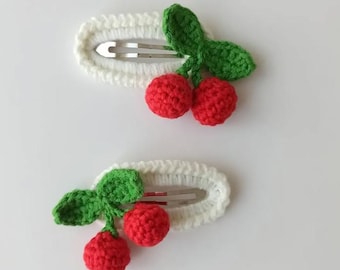 Puede incluir: Dos pinzas para el pelo de ganchillo blanco, cada una adornada con un grupo de cerezas rojas de ganchillo y hojas verdes. Las pinzas tienen un cierre de metal plateado. Los accesorios para el pelo están sobre un fondo blanco.