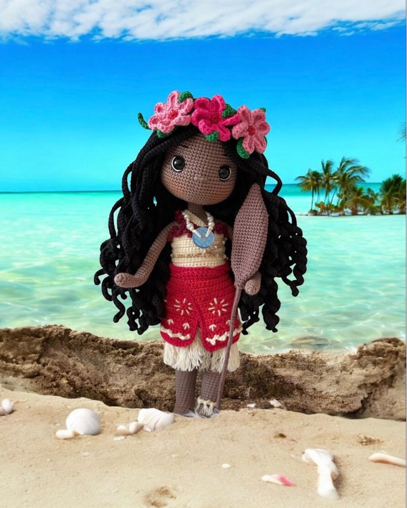May include: Crocheted doll with black hair, wearing a red skirt with white fringe and a floral crown. The doll is holding a wooden paddle and a necklace. The background shows a beach with blue water and a clear sky.