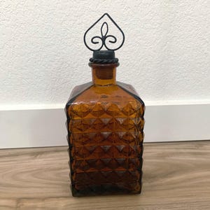 May include: Amber-colored glass decanter with a geometric diamond pattern. The bottle has a black metal stopper with a decorative heart-shaped design. The decanter is a decorative item for home decor.