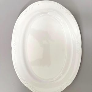 May include: A glossy white oval serving platter featuring a smooth, recessed centre and a raised, decorative rim with subtle, repeating embossed scrollwork patterns along the outer edge, providing a clean and uniform appearance for food service.