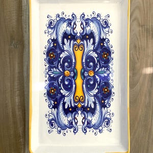 May include: Rectangular ceramic serving tray with a white base and a yellow border. The tray features a symmetrical design of blue and white stylized fish and floral motifs. The handles have a series of red and brown dots.