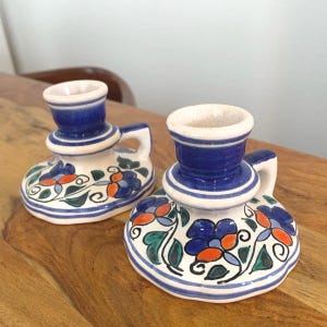 Könnte beinhalten: Zwei Kerzenhalter aus Keramik mit blau-weißen Streifen und floralen Designs. Die Kerzenhalter haben einen Griff und sind mit blauen, orangefarbenen und grünen Blumen und Blättern verziert. Die Kerzenhalter stehen auf einer Holzoberfläche.