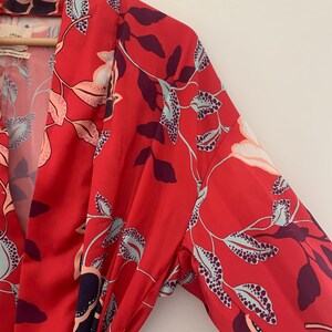 May include: A red floral print robe with long sleeves. The robe features a vibrant pattern of leaves and flowers in shades of purple, light blue, and peach. The fabric has a silky sheen, and the robe is partially open, revealing the interior.