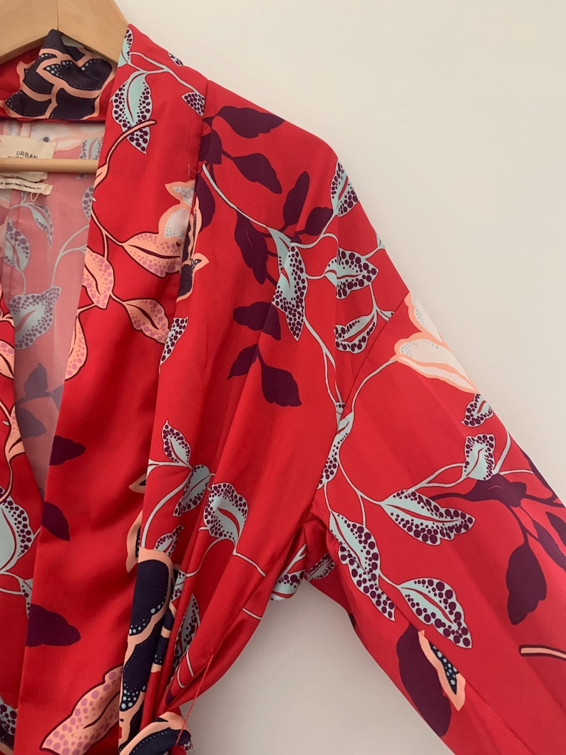 May include: A red floral print robe with long sleeves. The robe features a vibrant pattern of leaves and flowers in shades of purple, light blue, and peach. The fabric has a silky sheen, and the robe is partially open, revealing the interior.