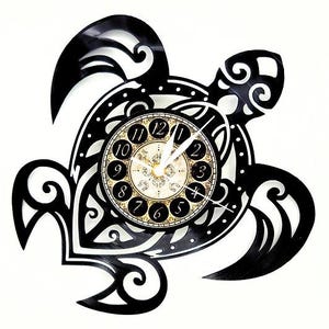 May include: Black turtle-shaped wall clock with a decorative design. The clock face features a cream-colored background with black numerals and gold accents. The clock hands are white.