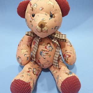 May include: A pink teddy bear with a brown and white checkered ribbon around its neck. The bear is made of fabric and has a pattern of children playing and animals on it. The bear has red and white polka dot feet.