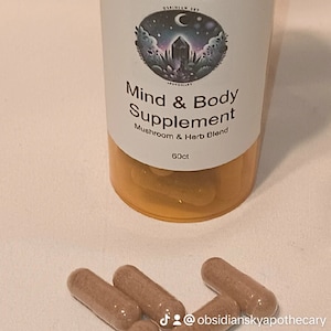 May include: An amber-colored prescription bottle with a white lid, containing capsules. The label reads "Mind & Body Supplement" with a mushroom and herb blend. Four capsules are scattered in front of the bottle. The text "60ct" is also visible.